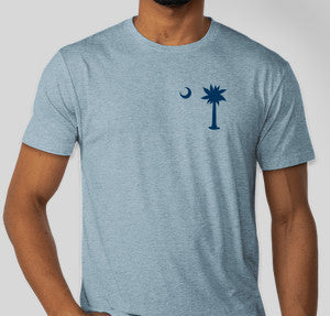 Person in a short sleeve light blue t-shirt with a navy palmetto tree and half moon on the front top left of the shirt. 